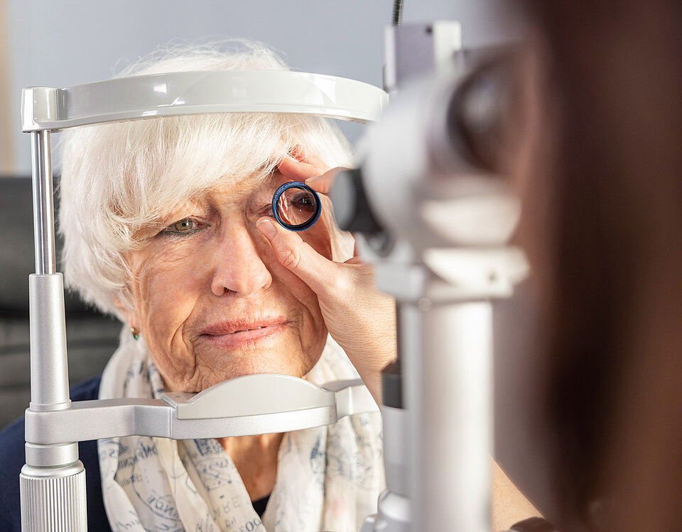 Augenuntersuchung bei Macuria Eine ältere Frau unterzieht sich einer Augenuntersuchung mit einem Gerät, das auf das Auge gerichtet wird, während eine Fachkraft hilft.